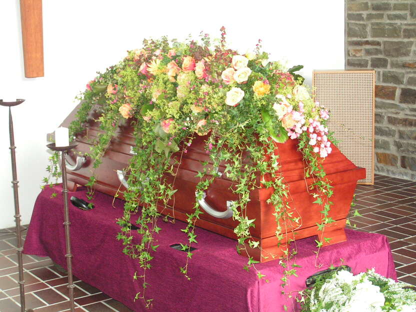 Holzsarg mit bunten Blumenarrangements und gr&uuml;ner Trauerfloristik auf einem lila Tischdecken in einer Trauerhalle.