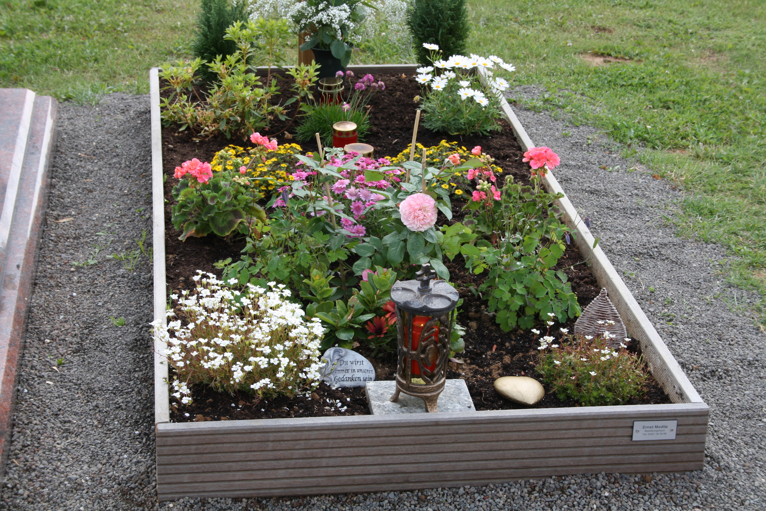 Blumenbeet mit bunten Blumen und einer Laterne auf einem Grab, umrandet von Kies auf einer gr&uuml;nen Wiese.