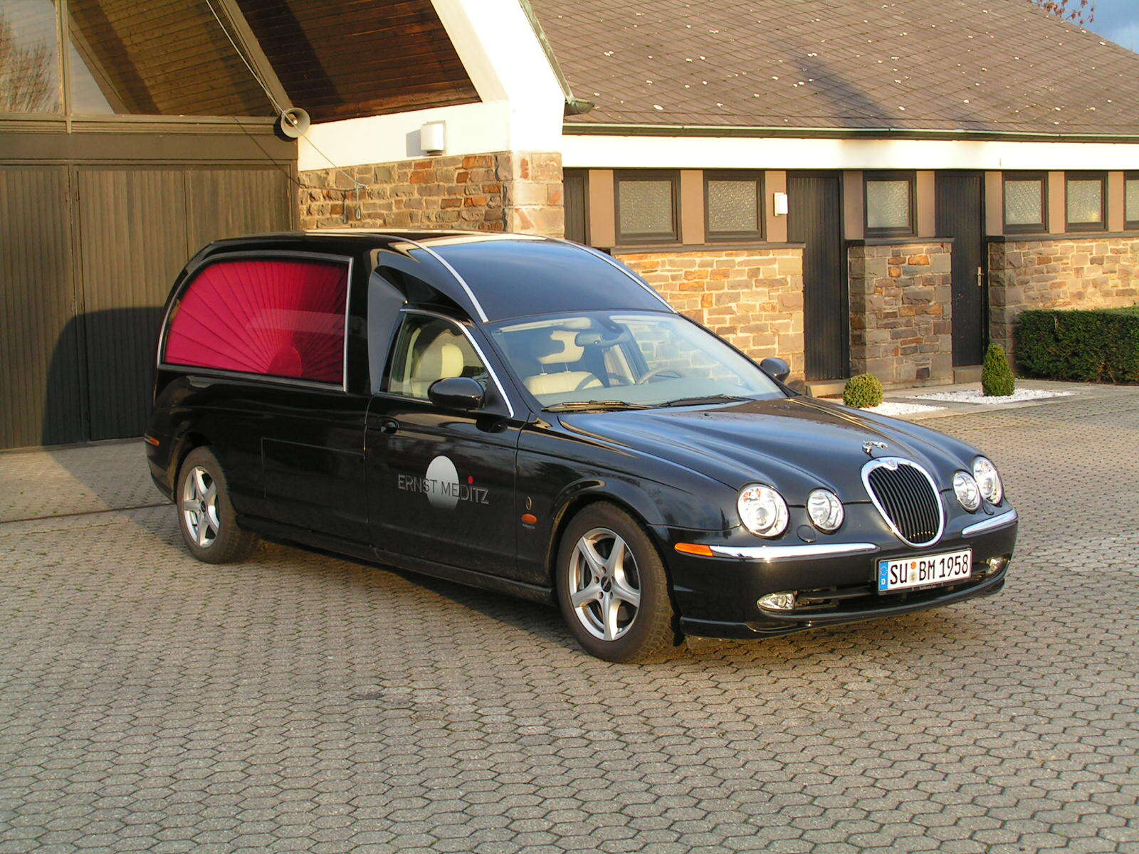 Schwarzer Leichenwagen mit pinken Details steht vor einem Geb&auml;ude auf einem gepflasterten Platz.