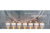Eine Reihe von brennenden Teelichtern auf einem Tisch vor einem grauen Wandhintergrund mit der Aufschrift &bdquo;Wir nehmen Abschied&ldquo;.