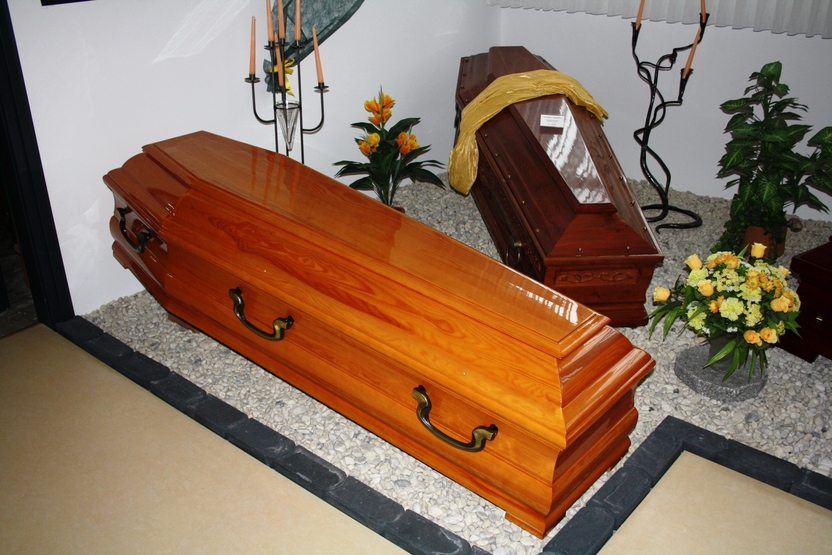 Holzfarbener Sarg auf Kiesboden, umgeben von Kerzen, Pflanzen und einem zweiten, dunkleren Sarg im Hintergrund.