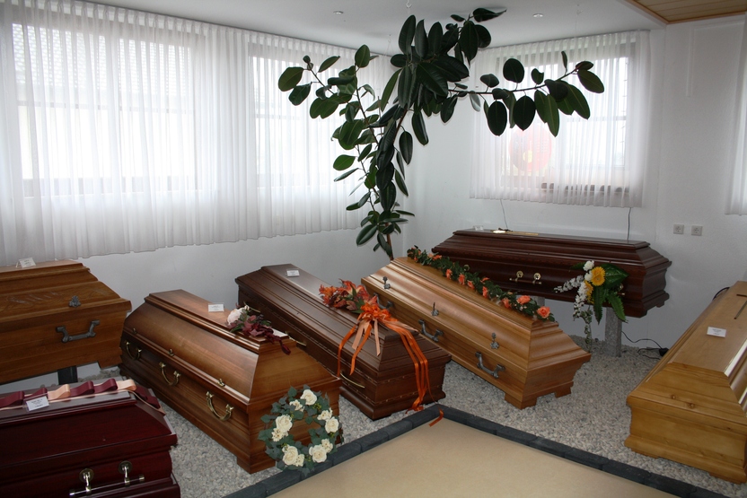 Mehrere klassische Holzs&auml;rge stehen in einem hellen Raum, dekoriert mit Blumen und einer Pflanze im Hintergrund.