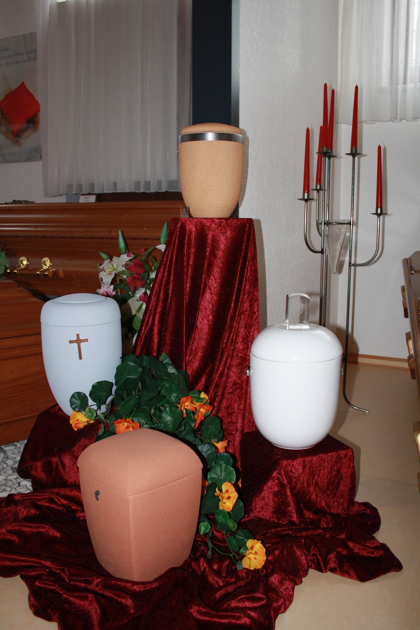 Drei Urnen auf einem rot drapierten Tuch in einem Raum, umgeben von Pflanzen und Kerzenst&auml;nder im Hintergrund.