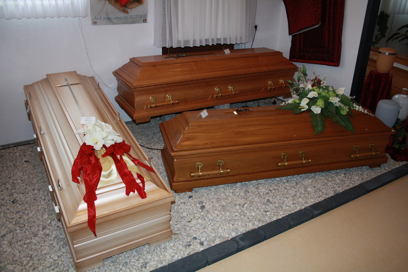 Drei S&auml;rge in einem Bestattungsinstitut, einer mit Blumenarrangement und rotem Band, umgeben von Kieselsteinen.