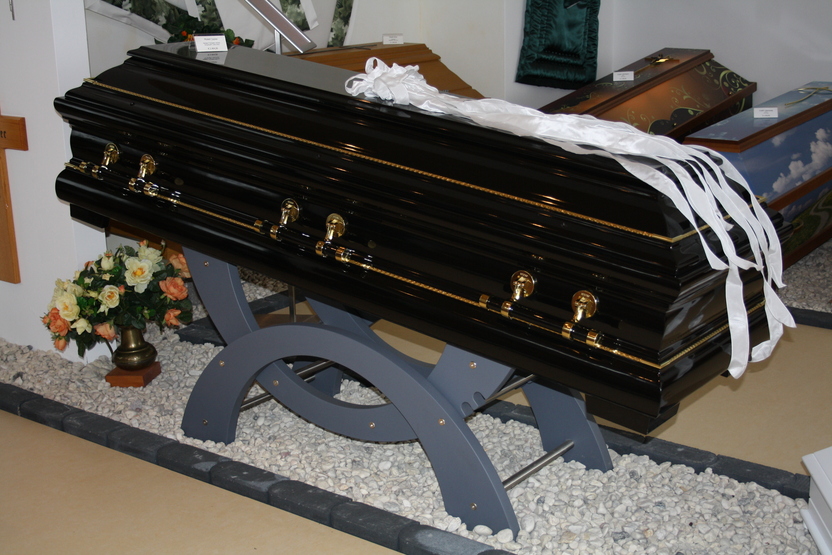 Schwarzer Sarg auf einem St&auml;nder aus Holz mit wei&szlig;en B&auml;ndern und Blumendekoration im Hintergrund.