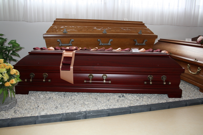 Drei S&auml;rge in einem Raum, der vordere in dunklem Holz mit einem Band verziert, Pflanzen und Steine im Vordergrund.