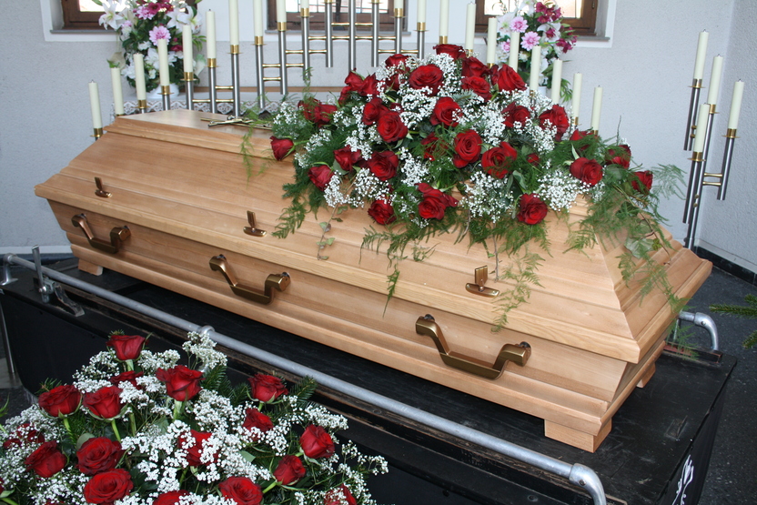 Holzlieferung eines Sarges mit roten Rosen und wei&szlig;en Blumenarrangements, umgeben von Kerzen und einer Trauerbank.