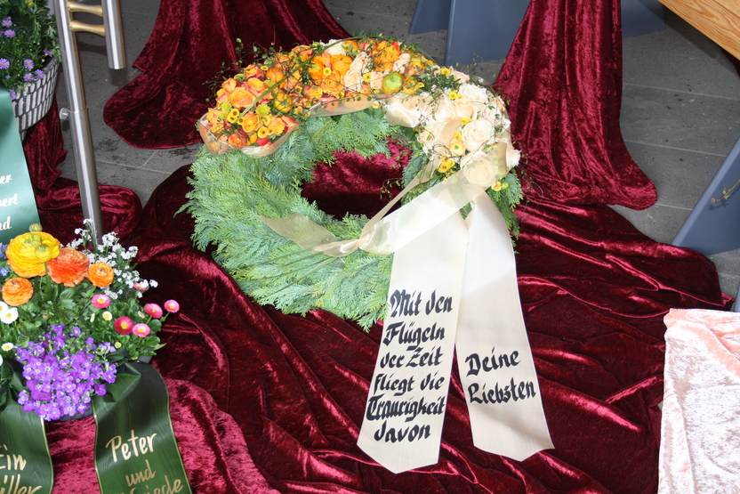 Blumenkranz mit bunten Blumen und einer Trauerschleife auf rotem Material, beschriftet mit: &bdquo;Mit den Fl&uuml;geln der Zeit...&ldquo;.