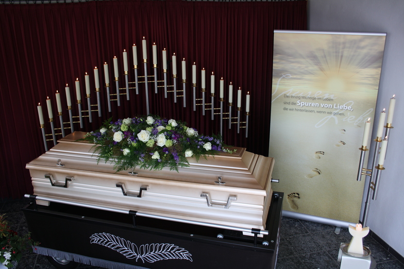 Ein offener Sarg mit Blumenarrangement in einem Raum, umgeben von Kerzen und einem aufgestellten Banner.