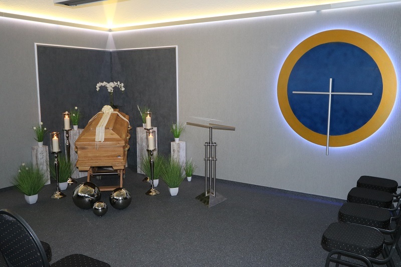 Holzkarton mit Blumen, Kerzen und Deko in einem trauernden Raum, hinter einem Podium mit minimalistischer Gestaltung.