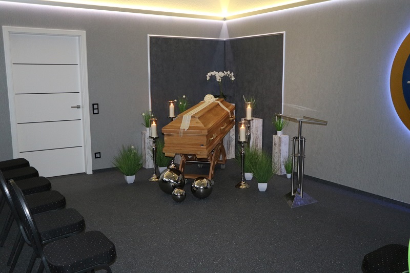 Holzsarg mit Blumen und Kerzen in einem schlicht gestalteten Trauerraum, umgeben von St&uuml;hlen und dekorativen Elementen.