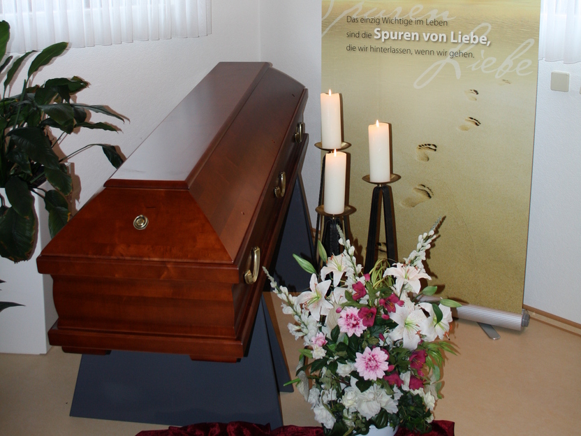 Holzkiste auf einem Tisch mit Blumenarrangement und Kerzen, hinterlegt von einem Banner mit einem einf&uuml;hlsamen Spruch.