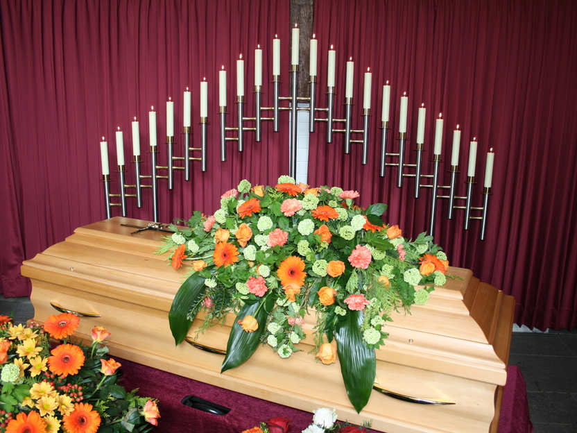 Holzschrein mit Blumenarrangement in leuchtenden Farben und Kerzen im Hintergrund, eingerahmt von roten Vorh&auml;ngen.