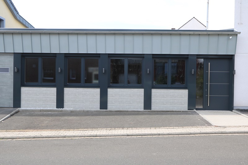 Fassade eines modernen Geb&auml;udes mit grauer Wand und gro&szlig;en Fenstern, seitlichem Eingang und einem flachen Dach.