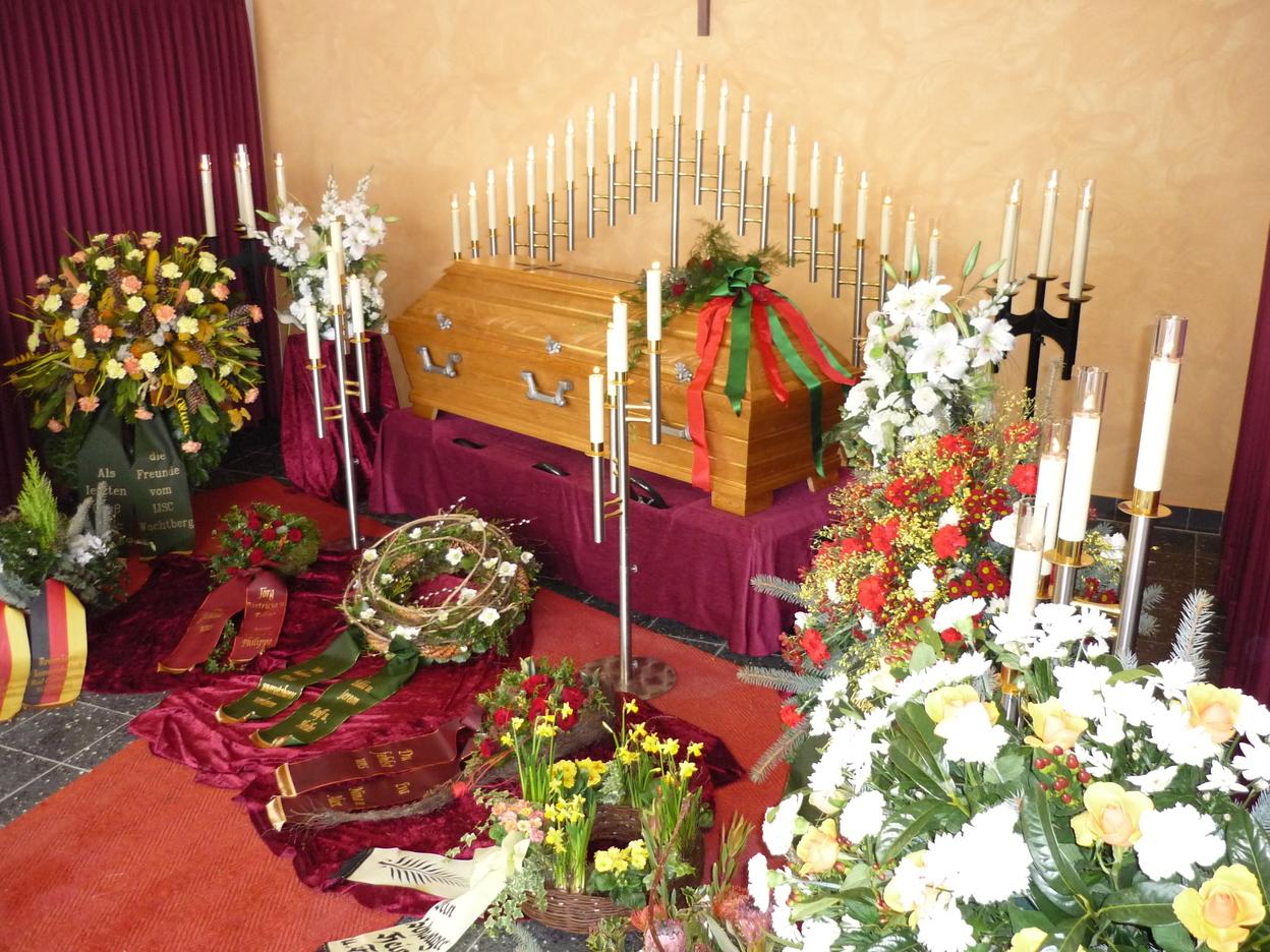 Blumenarrangements und Kerzen um einen aufgebahrten Sarg in einem festlich dekorierten Raum.