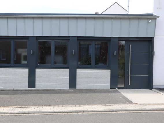 Fassade eines modernen Geb&auml;udes mit grauer Wand und gro&szlig;en Fenstern, seitlichem Eingang und einem flachen Dach.