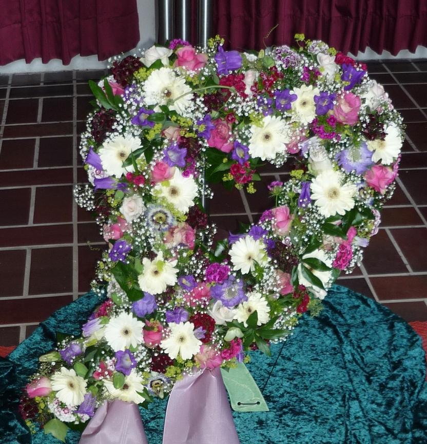 Herzf&ouml;rmiger Blumenkranz aus bunten Blumen auf gr&uuml;nem Tuch, mit einer lilafarbenen Schleife.