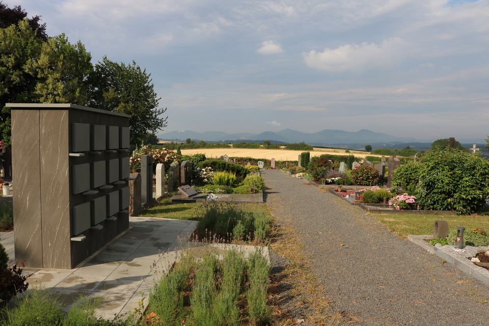 Friedhofsansicht mit Grabsteinen, Weg und Pflanzen, im Hintergrund Berge und ein bew&ouml;lkter Himmel.