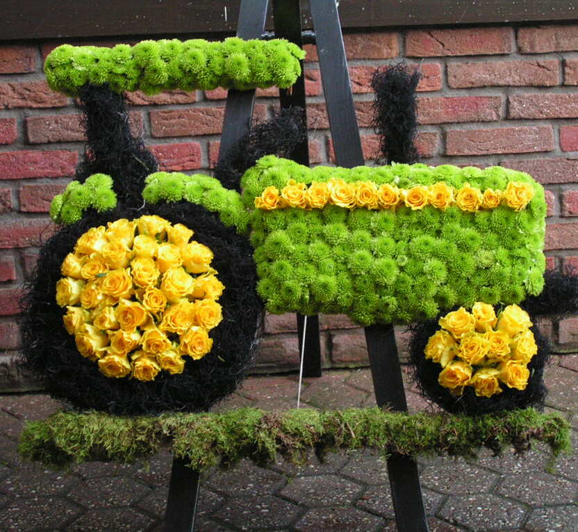Blumenarrangement in Form eines Traktors, dekoriert mit gelben und gr&uuml;nen Blumen auf einer Holzunterlage.