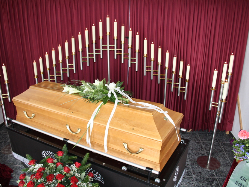Holzk coffin mit Blumenarrangement, umgeben von Kerzen und rotem Vorhang in einem Trauerraum.