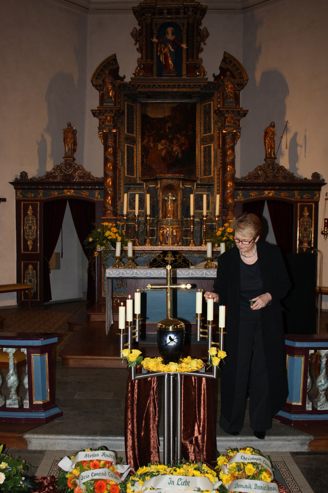 Frau in Schwarz steht vor einem Altar mit Kerzen und Trauerkr&auml;nzen in einer Kirche.
