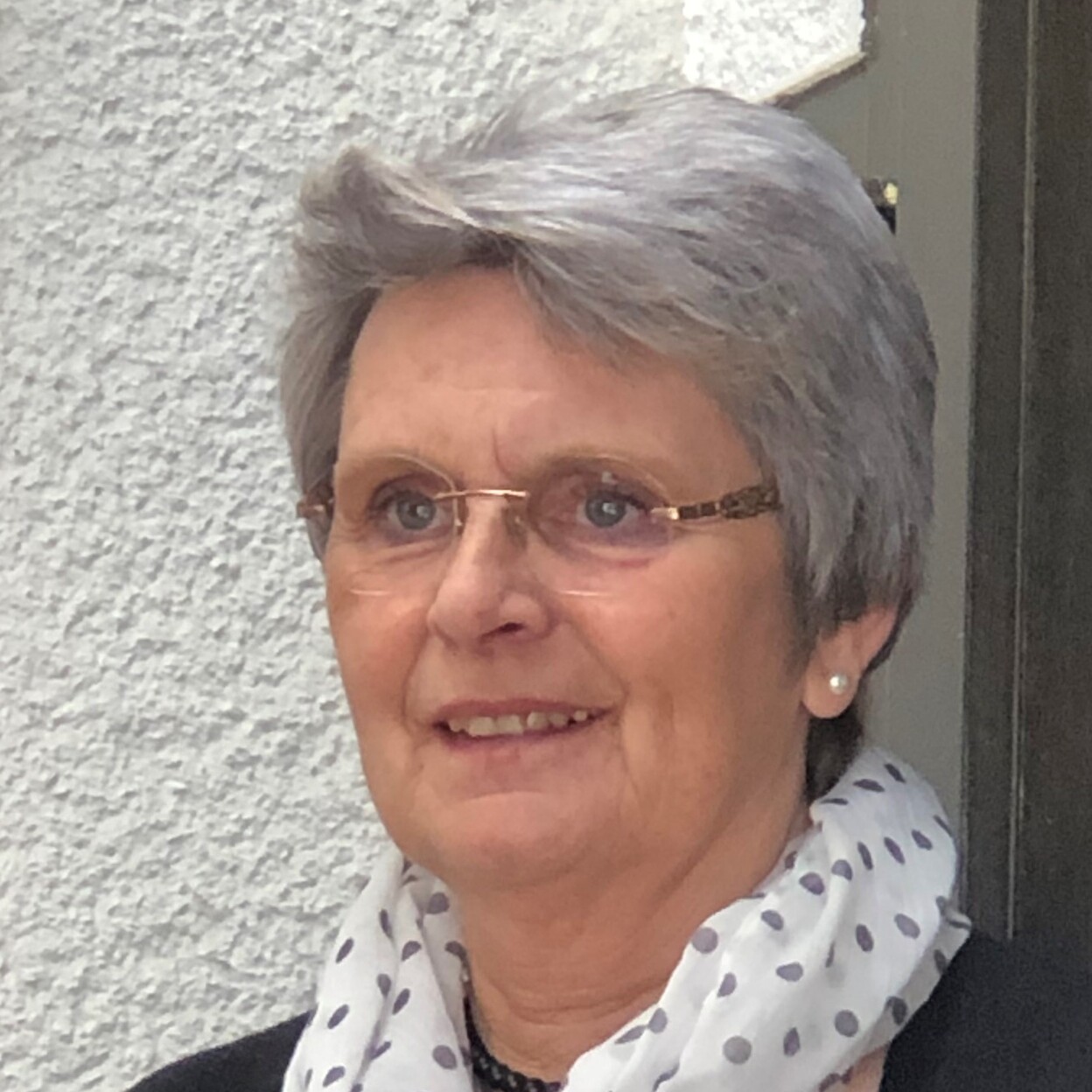 Portrait einer l&auml;chelnden Frau mit grauen Haaren, Brille und gepunktetem Schal vor einer hellen Wand.