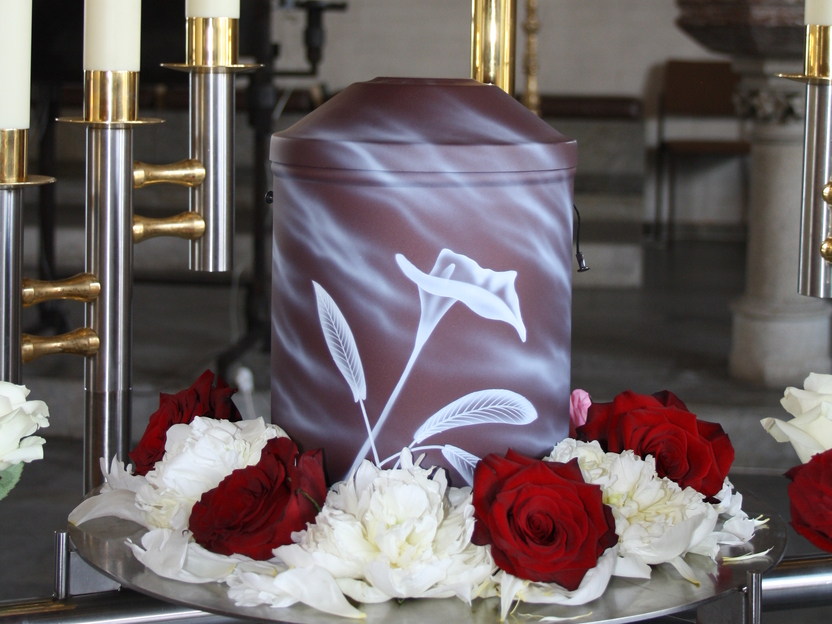 Urne mit floralem Motiv, umgeben von roten und wei&szlig;en Rosen, steht unter einem goldenen Kreuz in einer Kirche.