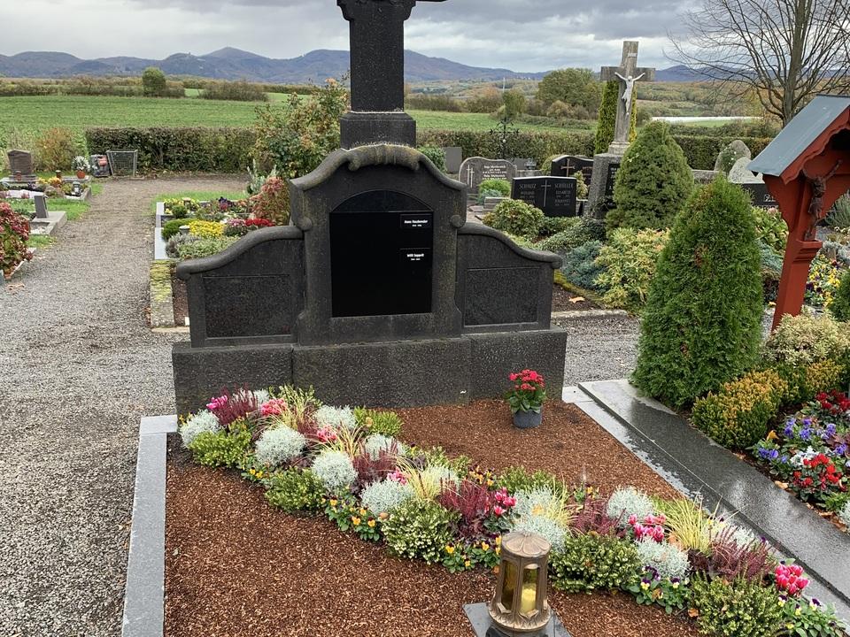 Schwarzes Grabkreuz auf einem gepflegten Grab mit bunten Blumen und einer Laterne in einer l&auml;ndlichen Umgebung.