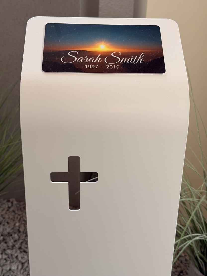 Wei&szlig;er Gedenkstein mit einem Foto und dem Namen &bdquo;Sarah Smith&ldquo; sowie den Jahren 1997 bis 2019, neben einer Kreuzgravur.