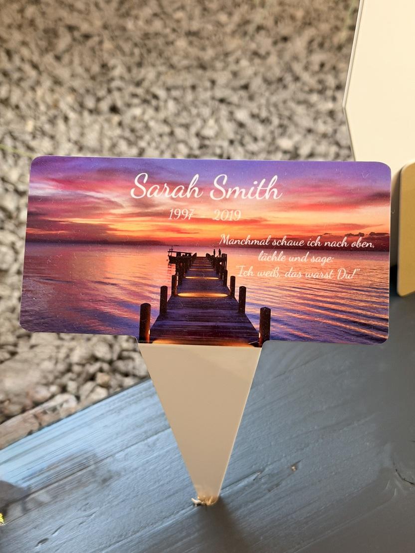 Gedenktafel f&uuml;r Sarah Smith mit Daten und einem Zitat, vor einem Sonnenuntergang &uuml;ber einem See, auf einem Tisch.