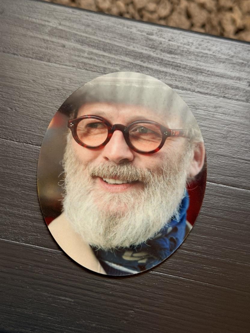 Ovales Foto eines l&auml;chelnden Mannes mit Brille und grauem Bart, auf einem dunklen Tisch liegend.