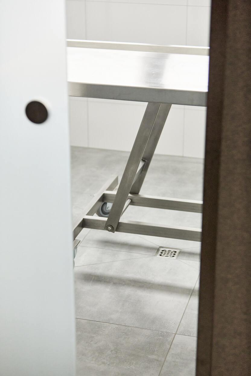 Blick durch eine Tür auf einen Metalltisch mit Rollen in einem gefliesten Raum.