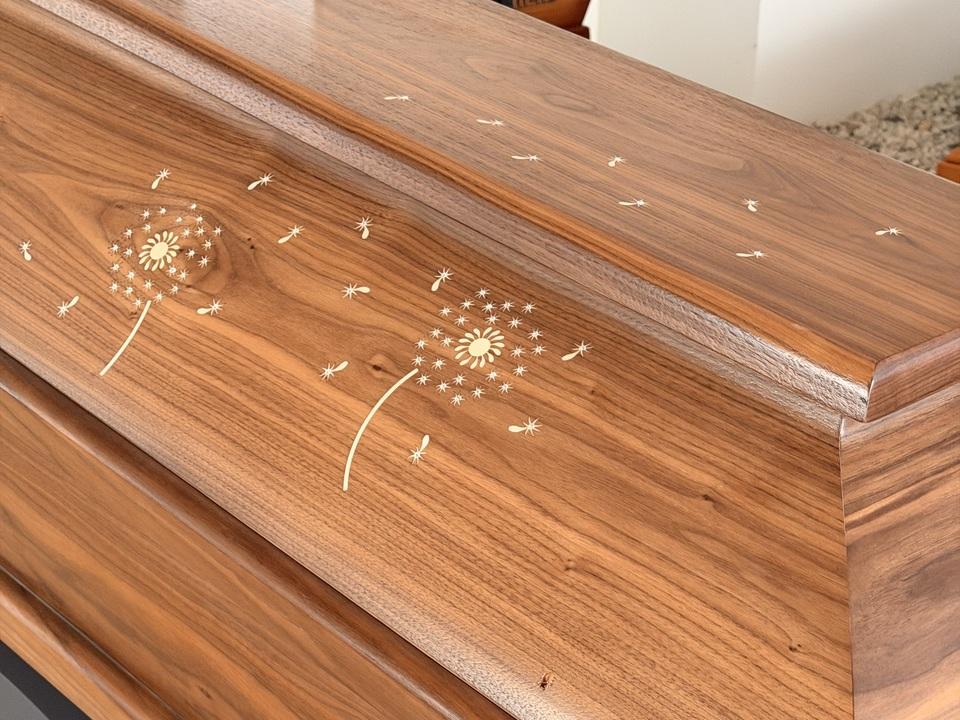 Holzurne mit goldenen Pusteblumen-Motiven und einem Griff, auf einem hellen, steinigen Untergrund platziert.