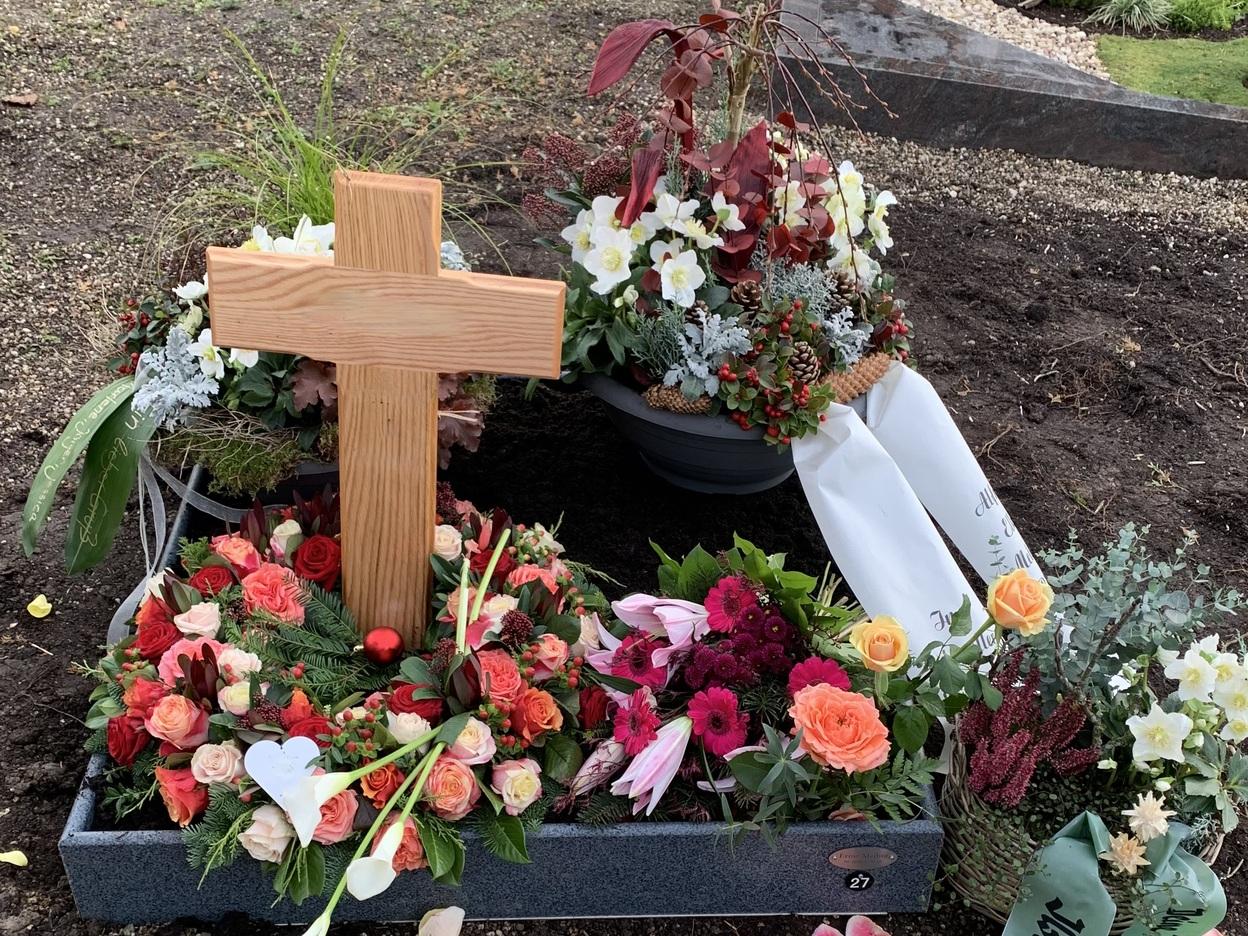 Grab mit einem h&ouml;lzernen Kreuz, umgeben von bunten Blumen und Gestecken, auf einem gepflegten Friedhof.