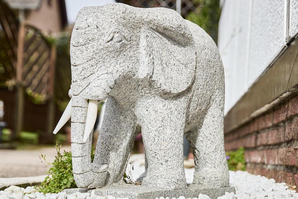 Steinskulptur eines Elefanten steht auf weißem Kies im Garten. Pflanzen und eine Hauswand im Hintergrund.