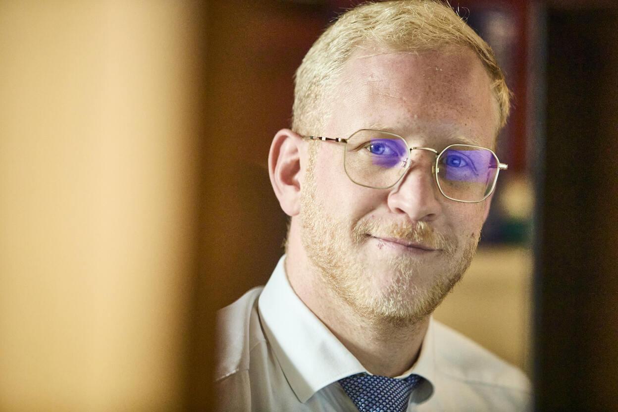 Ein Mann mit Brille und blondem Haar lächelt leicht, während er hinter einem Schreibtisch sitzt.