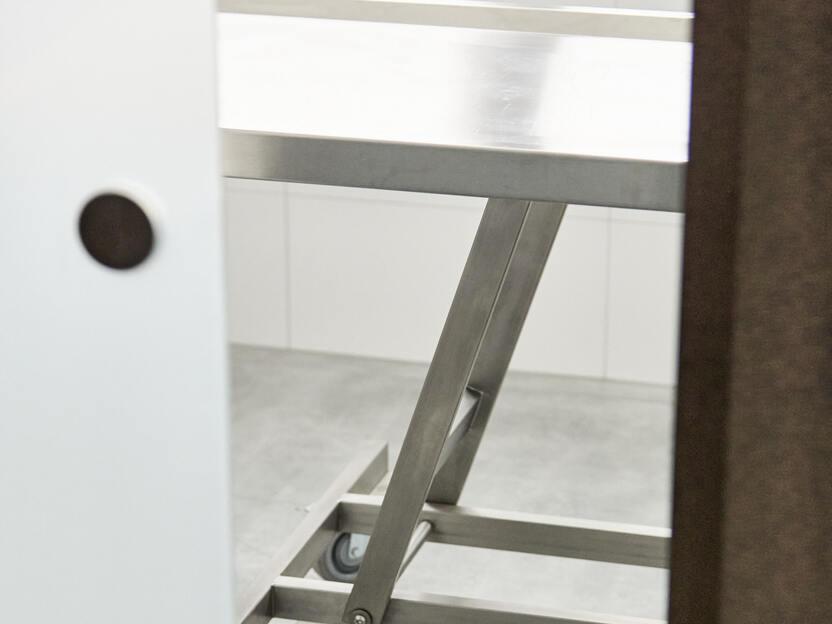 Blick durch eine T&uuml;r auf einen Metalltisch mit Rollen in einem gefliesten Raum.