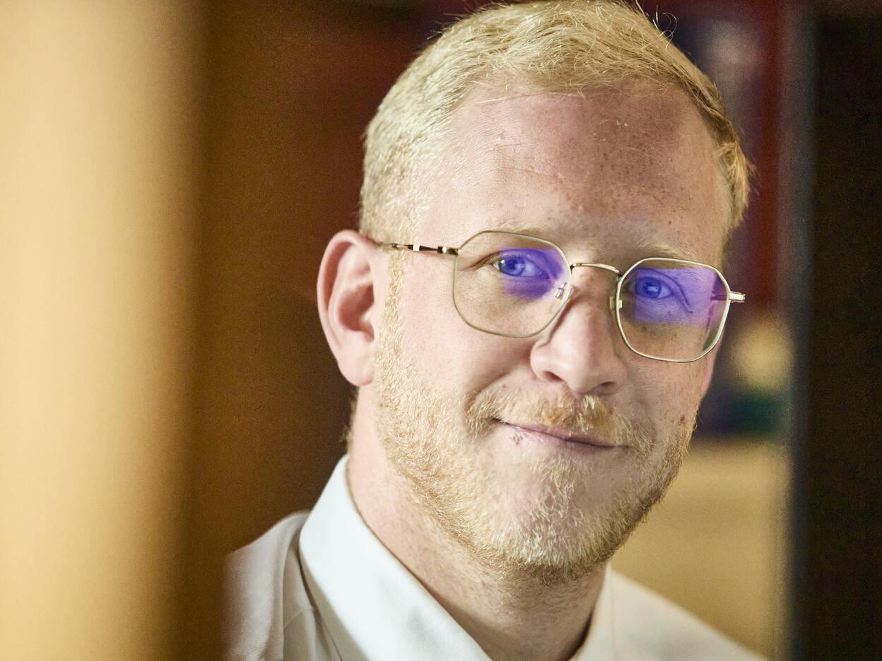 Ein Mann mit Brille und blondem Haar l&auml;chelt leicht, w&auml;hrend er hinter einem Schreibtisch sitzt.