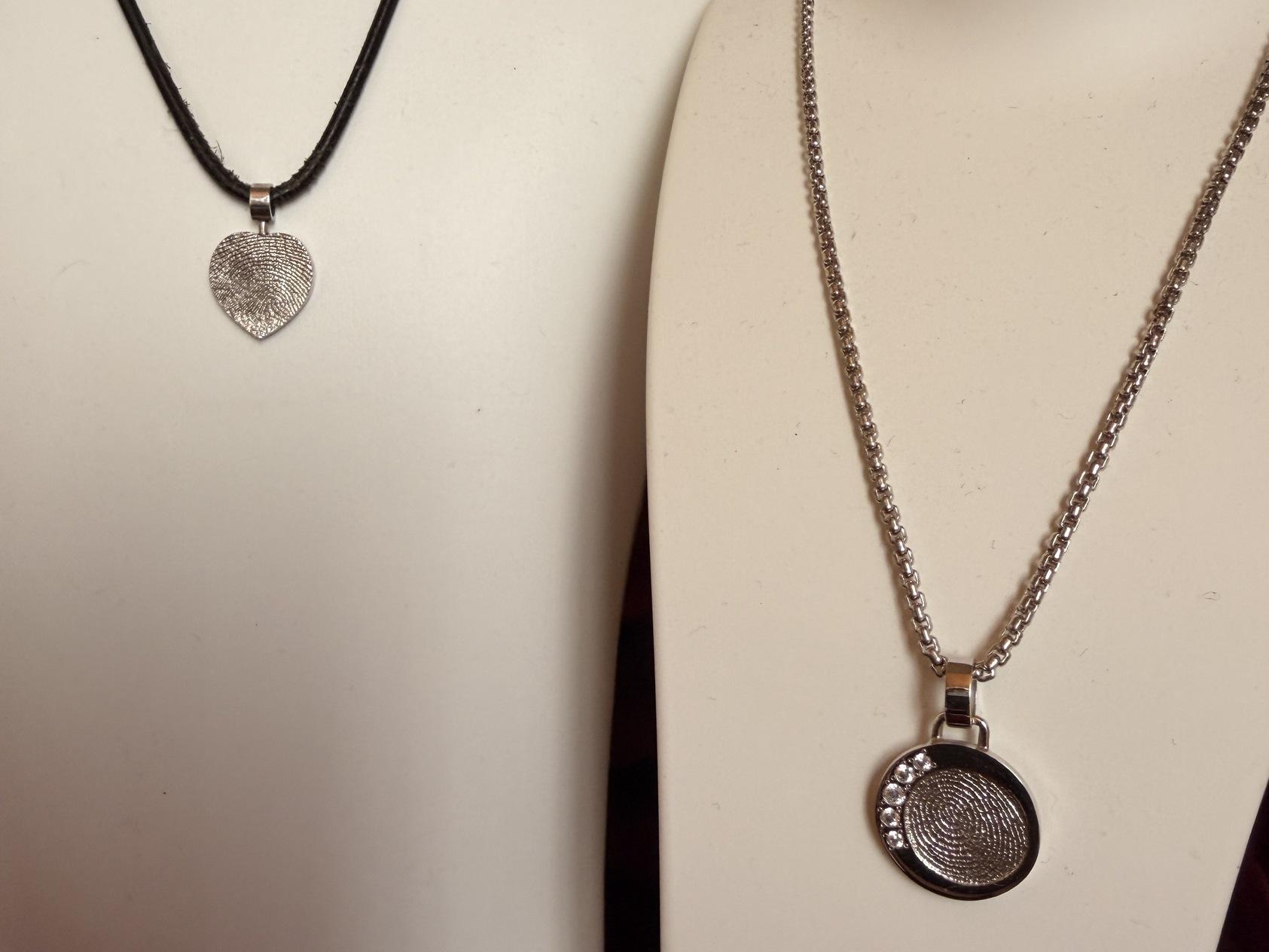 Zwei Halsketten an Schmuckst&auml;ndern: Eine schwarze mit herzf&ouml;rmigem Anh&auml;nger, eine silberne mit runder, gemusteter Medaille.