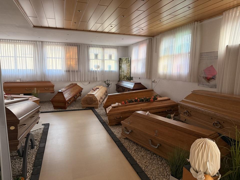 Ausstellung von mehreren S&auml;rgen in einem hellen Raume mit gro&szlig;en Fenstern und dekorativen Elementen.