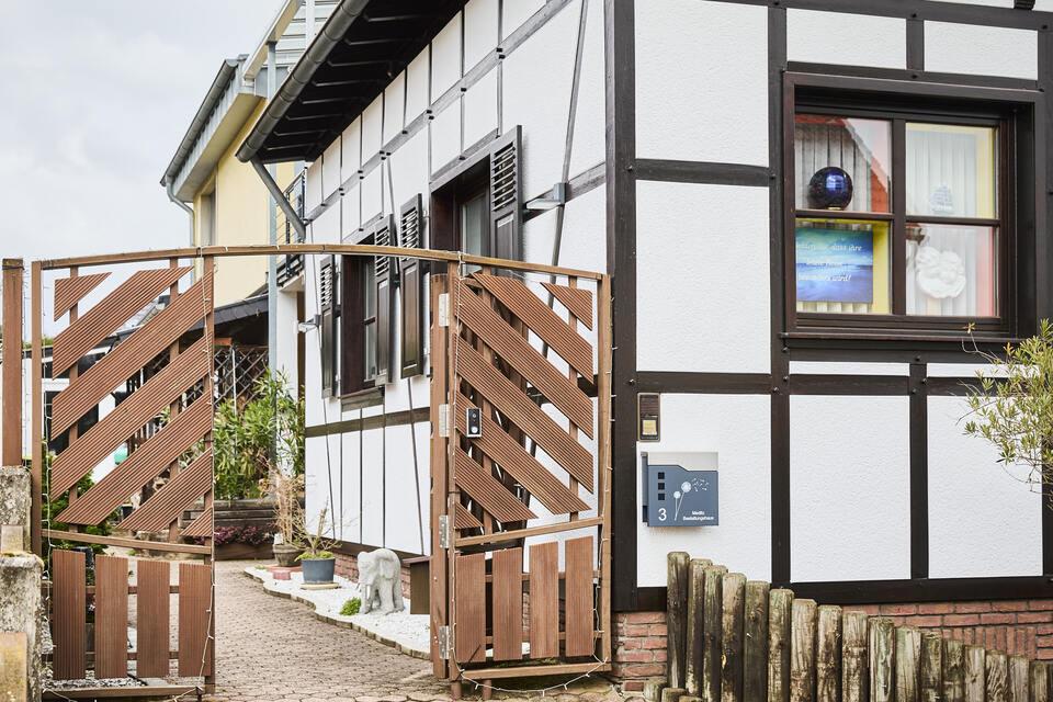 Fachwerkhaus mit offenem Holztor und buntem Fenster, umgeben von einem gepflasterten Weg und einem Garten.