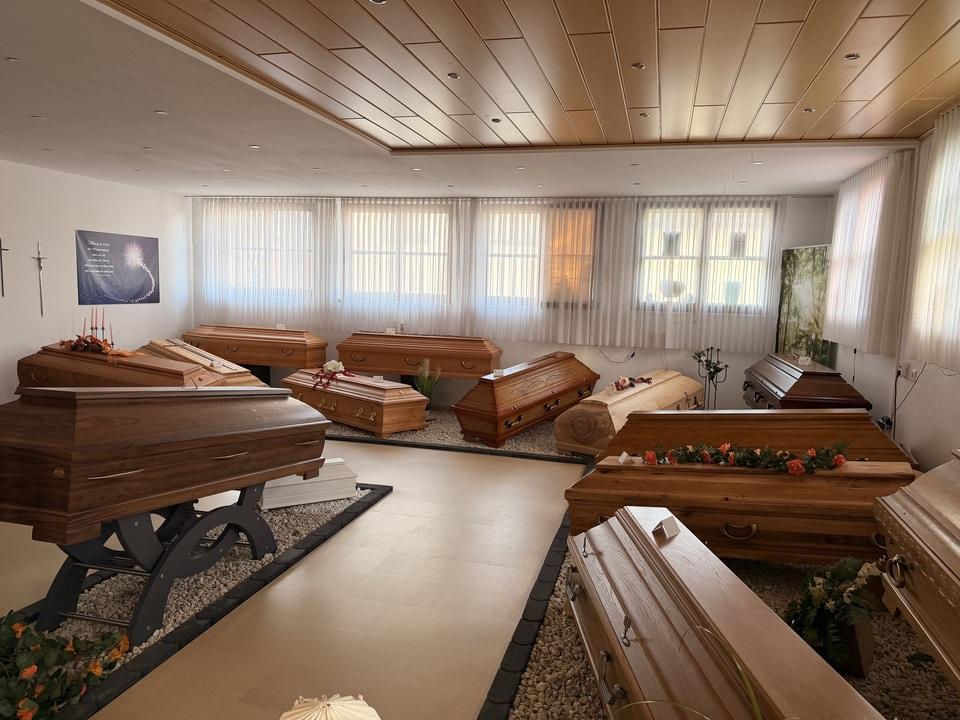 Raum mit mehreren Holzkirchen, dekoriert mit Blumen, unter Fensterfront mit Gardinen. Licht f&auml;llt sanft auf die S&auml;rge.