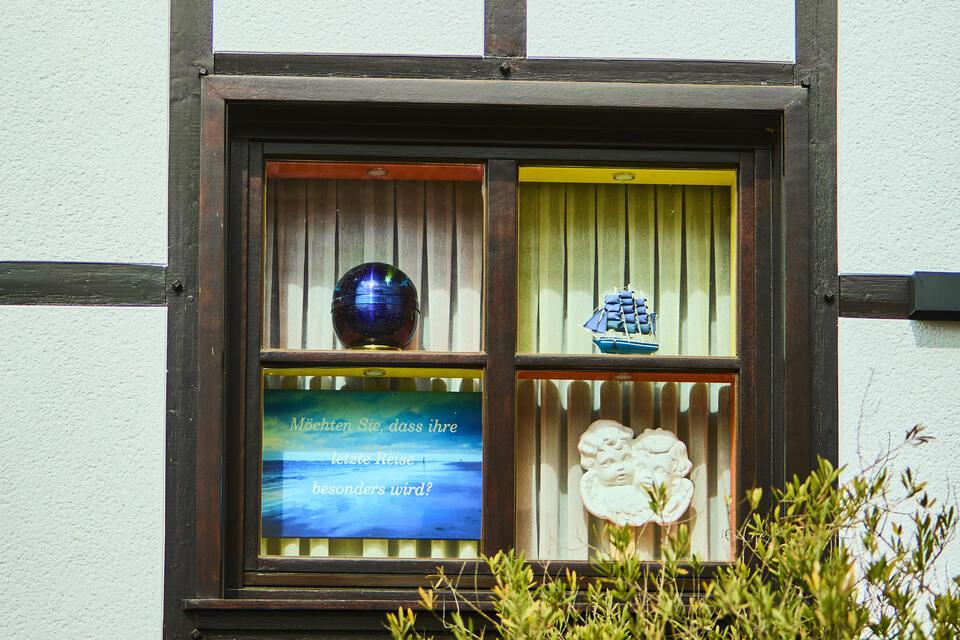 Blick auf ein zweigeteiltes Fenster mit bunten Deko-Objekten und einem Spruch auf einem der Gardinen.