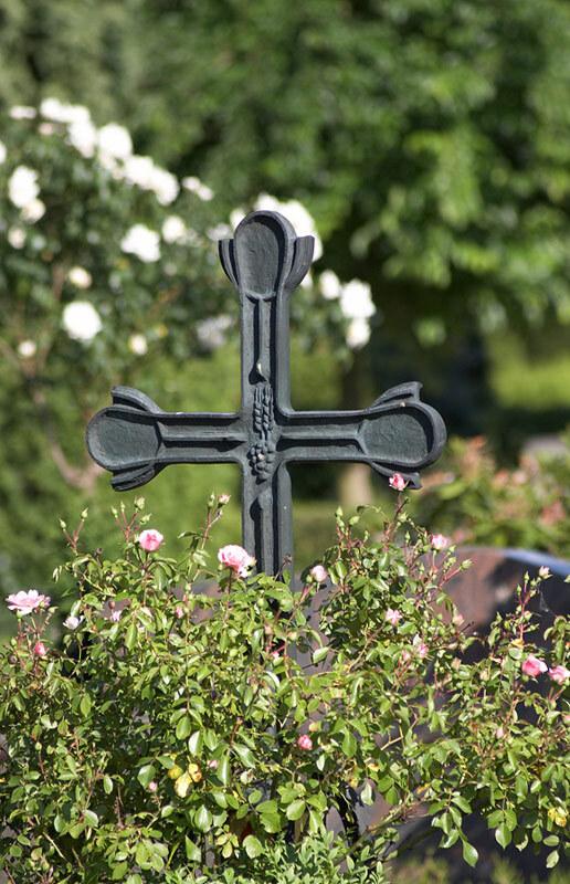Schwarzes Kreuz umgeben von bl&uuml;henden Rosen auf einem friedlichen Hintergrund mit B&auml;umen.