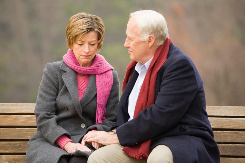 Zwei Personen sitzen auf einer Bank, halten sich an den H&auml;nden und blicken nachdenklich in die Ferne.