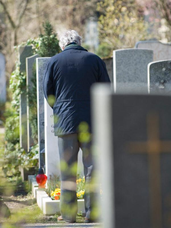 &Auml;lterer Mann steht vor einer Grabst&auml;tte auf einem Friedhof, umgeben von Grabsteinen und Blumen.