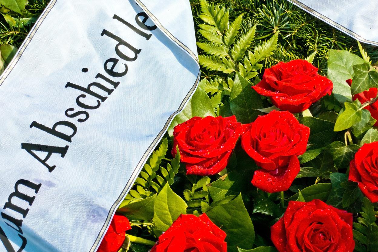 Rote Rosen mit Wassertropfen und einem weißen Band mit der Aufschrift „Zum Abschied“ auf grünem Hintergrund.