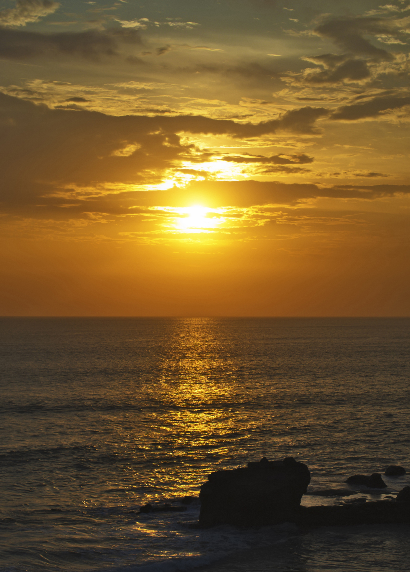 Sonnenuntergang über dem Meer, das sanftes goldenes Licht reflektiert, mit silhouettierten Felsen im Vordergrund.