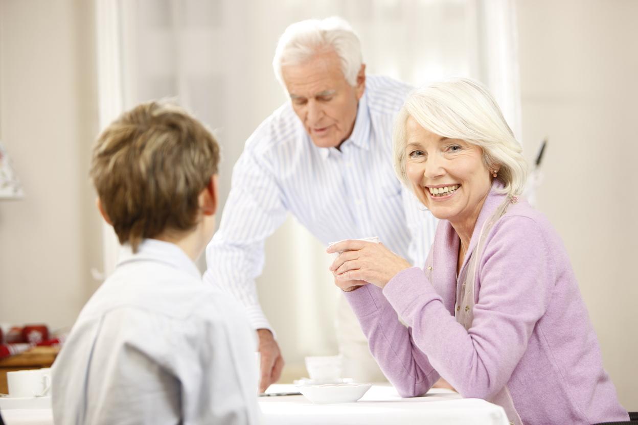 Lächelnde ältere Frau sitzt am Tisch, während ein Mann im Hintergrund Getränke serviert, ein Junge schaut sie an.