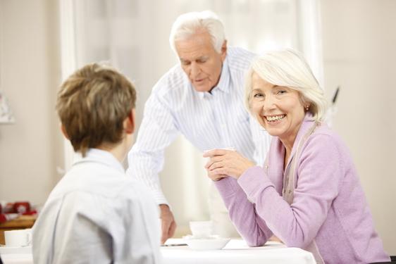 Lächelnde ältere Frau sitzt am Tisch, während ein Mann im Hintergrund Getränke serviert, ein Junge schaut sie an.